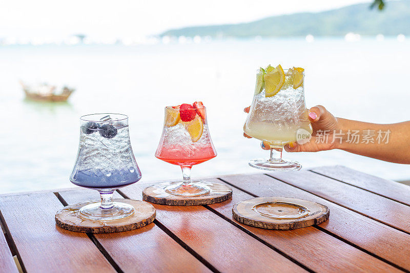 一只手拿着一杯意大利柠檬苏打饮料:草莓，橘子和蓝色夏威夷顶部混合水果在一个鸡尾酒杯，服务在木玻璃垫子上，在木桌子上，海滩背景，正面视图，水平