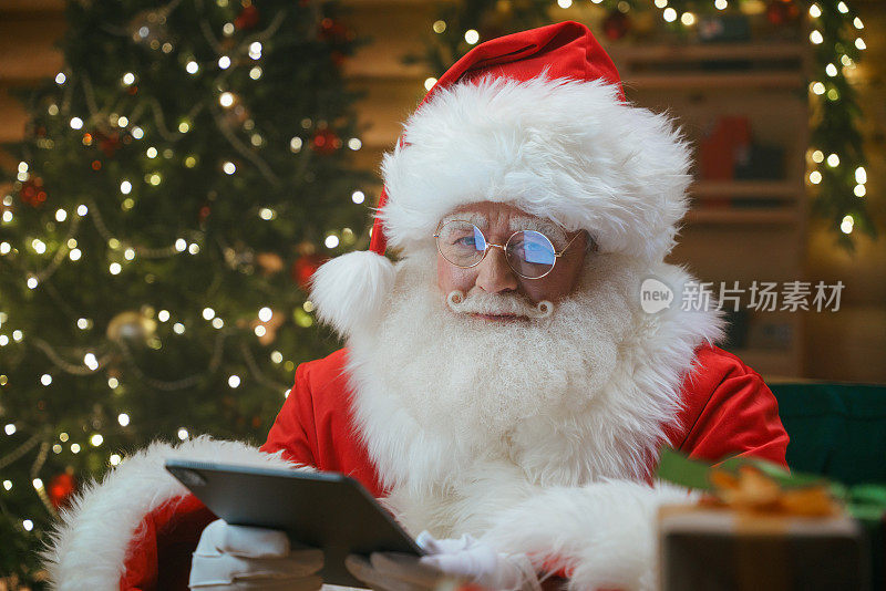 圣诞老人手持数码平板电脑，坐在装饰有圣诞树的小木屋里的一张桌子前