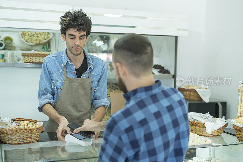 年轻的男面包房员工和顾客用终端和智能手机进行非接触式支付