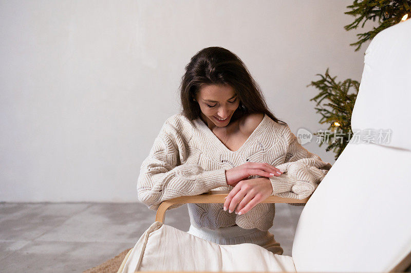 年轻美丽的女人模型在白色针织休闲毛衣坐在客厅附近的圣诞树，圣诞节和新年的概念