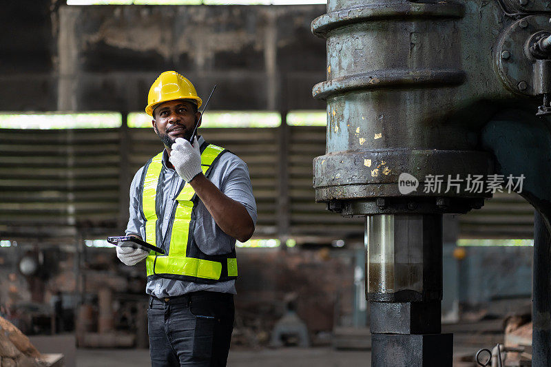 非裔美国男性工程师，在工业工厂检查或维护重金属机械。男技术员佩戴安全帽，制服在工厂内使用无线电通信工作