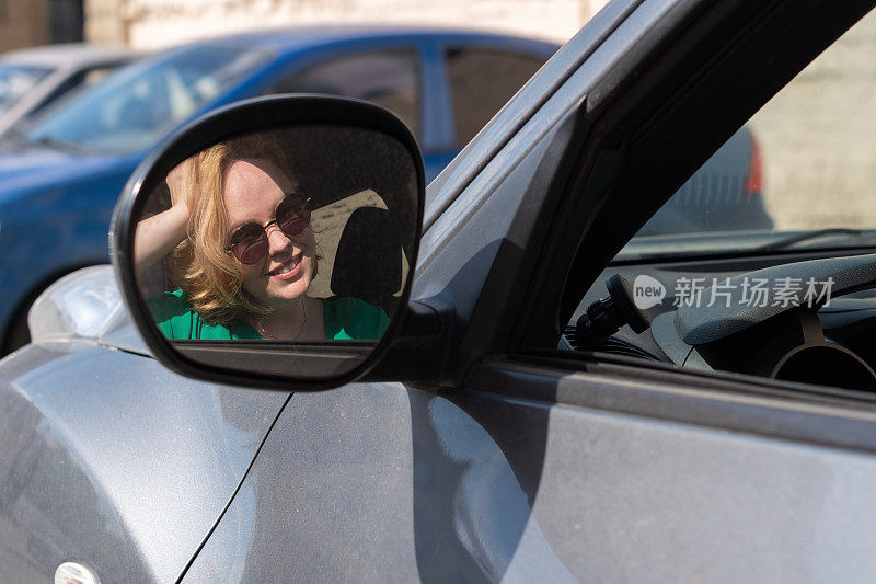 镜子里的倒影，戴着墨镜的微笑女孩坐在停车场的一辆车里向外看。