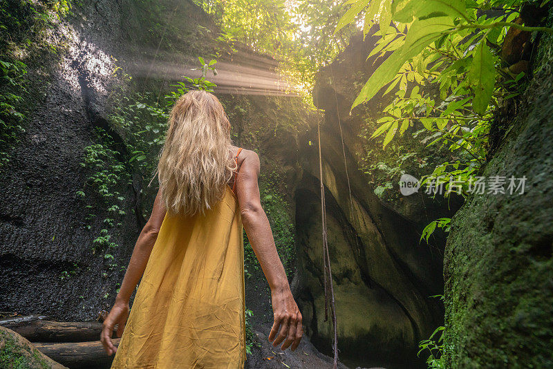 穿着黄色衣服的女孩站在热带雨林的洞穴里，仰望着从岩石上射下来的壮观的阳光。人们旅行的奇妙欲望自然的概念