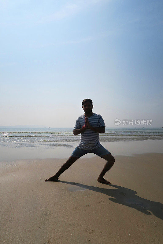 印度男子练习侧弓步(Skandasana)瑜伽姿势在潮湿的沙滩上，水的边缘，Palolem海滩，果阿，印度