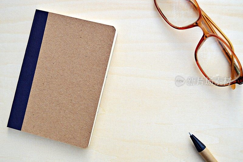 一张水平的照片，一张棕色的硬纸板装订的记事本，一只棕色和黑色的钢笔和眼镜，放在一个木制的米色背景上，很有美感。