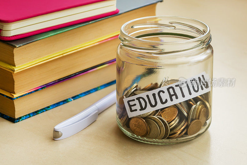 白色圆珠笔旁边放着很多硬币和教育字的玻璃罐子，桌上放着几本书。省钱为教育理念。