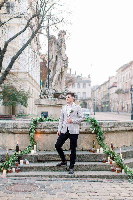婚礼前，穿着灰色夹克、面带微笑的新郎站在老城区纪念碑的石阶上，在户外摆姿势