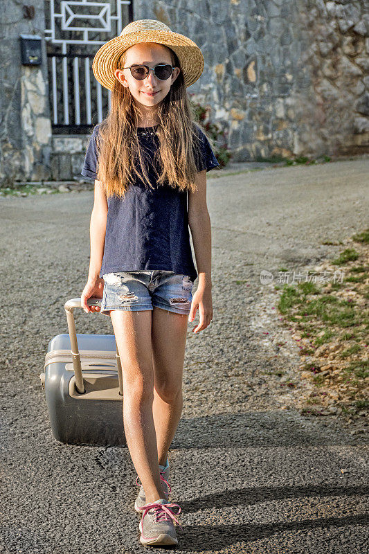 年轻的女孩走在乡村街道上的暑假拉着手提箱