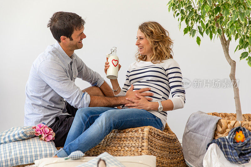 快乐的妻子和丈夫在一起大笑，同时喝牛奶和抱着怀孕的妻子的肚子-股票照片