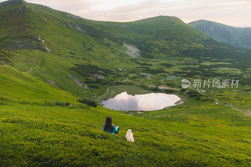 一个徒步旅行者和一只狗坐在草地上欣赏山上湖上美丽的日落