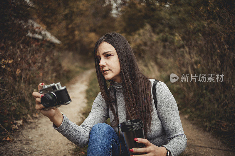 在大自然中摄影——快乐的年轻女子微笑着享受大自然，走过画着秋天颜色的森林，戴着复古的相机拍摄树叶