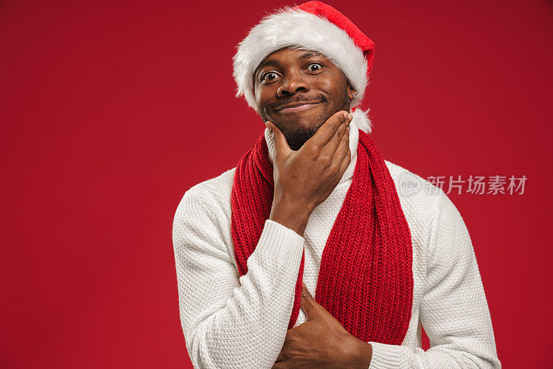 一个皮肤黝黑的男人，穿着羊毛衫，戴着圣诞帽，手拿下巴微笑着