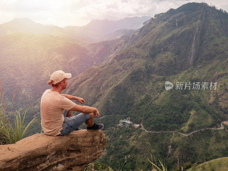 一个年轻人坐在斯里兰卡美丽山脉顶端的岩石上，欣赏着郁郁葱葱的风景