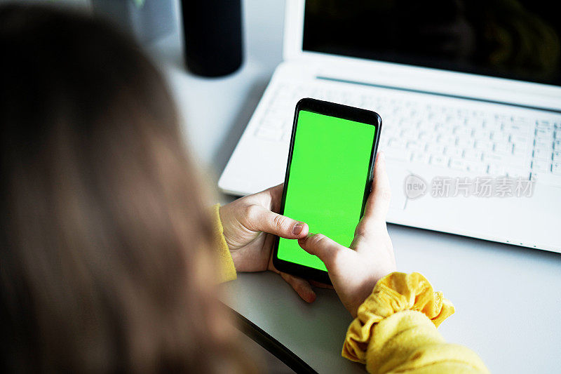 特写的十几岁的女孩手持智能手机与色度键绿色屏幕对白色笔记本电脑
