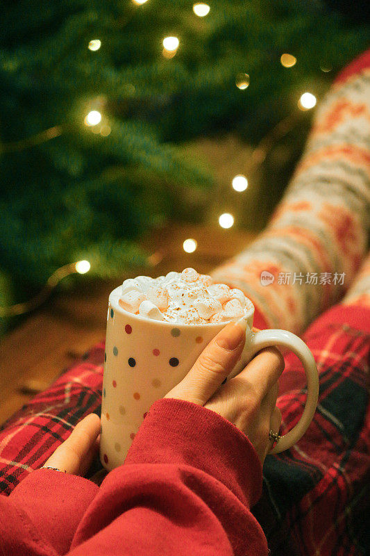 一个无法辨认的女人穿着红色格子睡衣，穿着圣诞主题的图案袜子，脚放在咖啡桌上，手里拿着斯波蒂杯里的热巧克力，上面有鲜奶油和迷你棉花糖，圣诞树上挂着发光的仙女灯