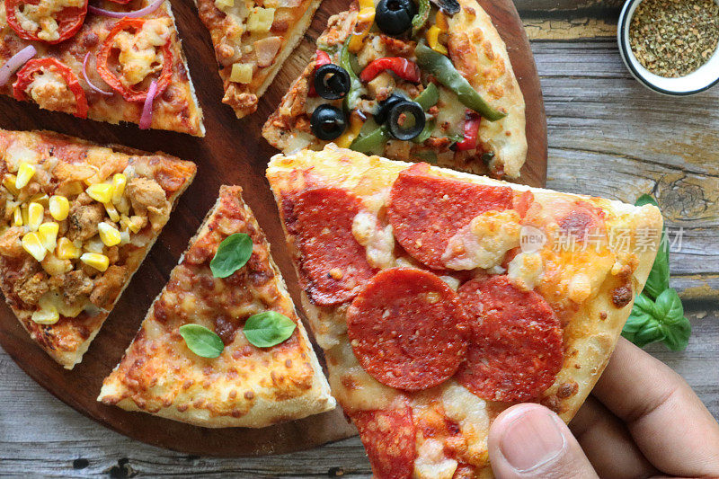 照片中，一个面目全非的人在挑选披萨片，各种配料，包括夏威夷披萨、羊乳酪、番茄片和红洋葱、鸡肉和甜玉米、意大利辣香肠、玛格丽塔披萨和圆形木砧板上的黑橄榄蔬菜