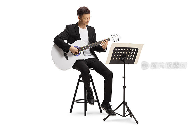 穿着黑色西装的年轻人坐在椅子上，用乐谱架演奏原声吉他