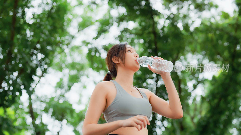 年轻的亚洲女性喝瓶装水，在她的锻炼期间休息。快乐的青春期女性在公园锻炼和休息休息喝水