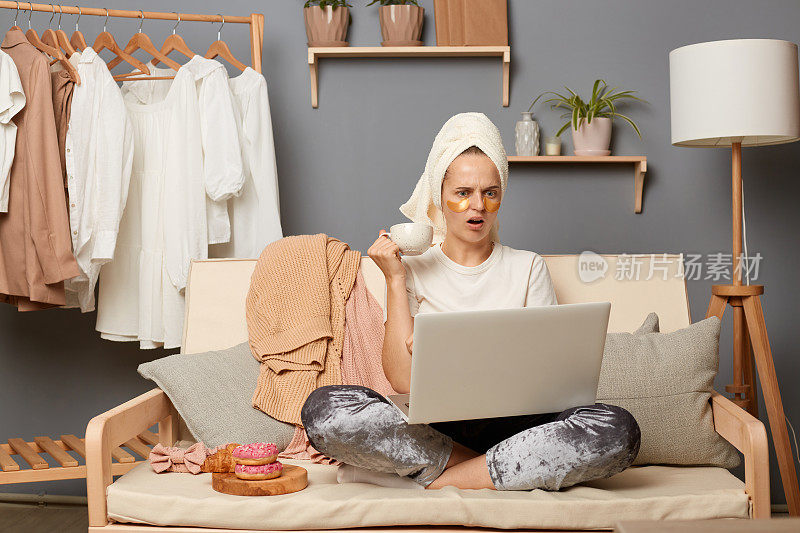 室内拍摄的震惊震惊的女人穿着白色t恤，灰色裤子，头上戴着毛巾，坐在咳嗽，膝盖上放着笔记本电脑，拿着一杯咖啡，阅读突发新闻。