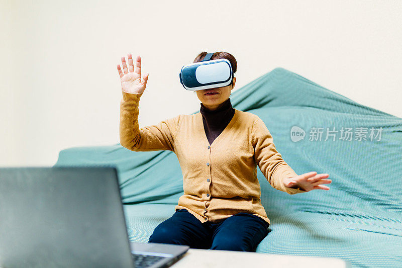 一位中年妇女戴着VR眼镜坐在家里的沙发上，用着笔记本电脑——科技与生活，健康的生活方式