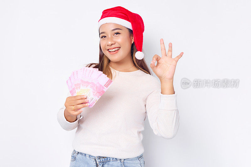 微笑的年轻亚洲女子20岁戴着圣诞帽，上面有现金、印尼盾和白色背景上的okay标志。2023年圣诞快乐
