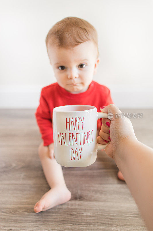 一个14个月大的可爱男婴光着脚，穿着写着“最小的情人节”的红毛衣和布尿布，坐在木制硬木地板上，他的妈妈在他面前举着一个写着“情人节快乐”的杯子