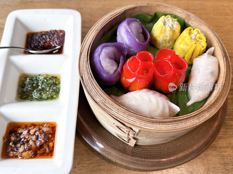 部分美食的特写图像，在竹制蒸笼里的五颜六色的鸡momos，内衬香蕉叶，除了蘸酱托盘，木纹背景，高架视图