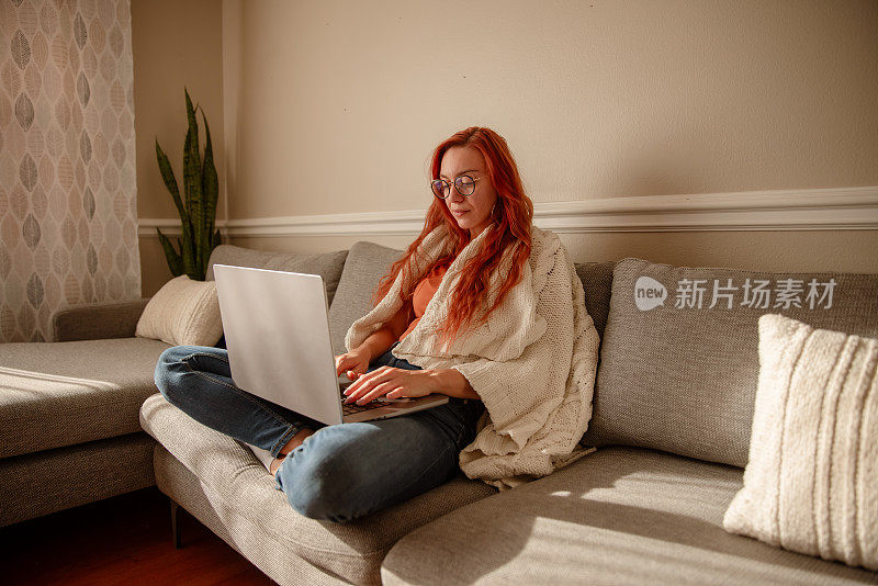 一位戴着眼镜、红发的年轻美女在家工作，同时处理多项任务。她裹着毯子坐在沙发上，膝上放着笔记本电脑。