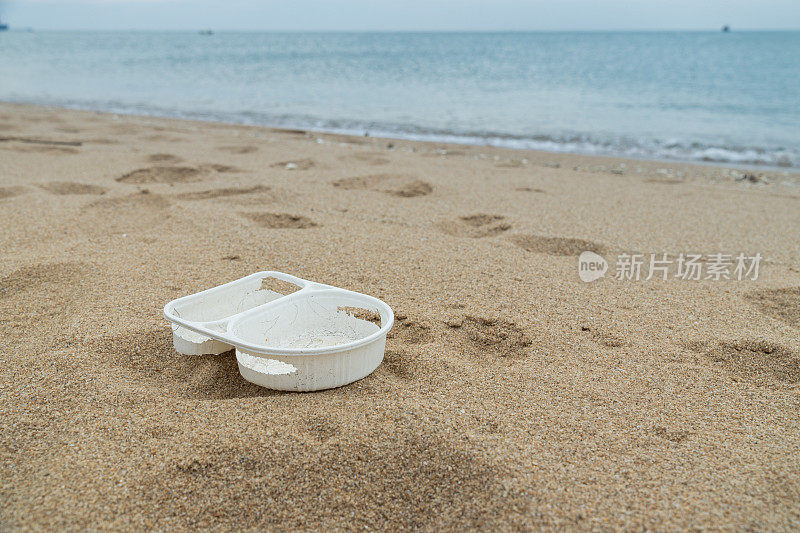 沙滩上白色塑料食品容器的特写镜头，背景是大海，显示塑料垃圾对环境的污染。