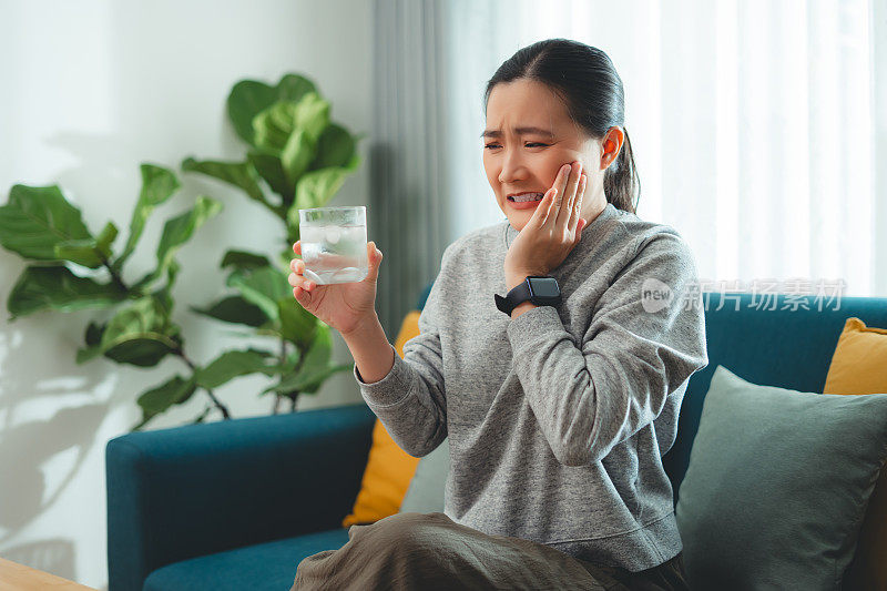 亚洲女性坐在客厅的沙发上，喝了凉水，摸了摸脸颊，就会感到牙齿敏感。