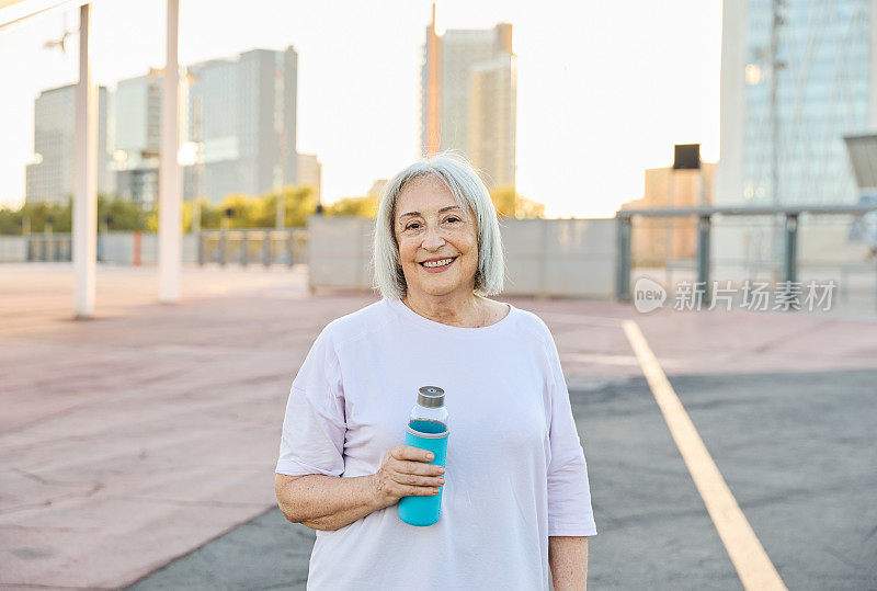 在运动场上，一位快乐的年长妇女站在那里，手里拿着水瓶，面带微笑