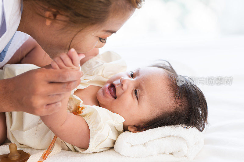 幸福的家庭。亚洲妈妈抱着她可爱的3个月大的白种人女婴的手，躺在家里的床上一起玩耍。可爱的婴儿看着妈妈，微笑着