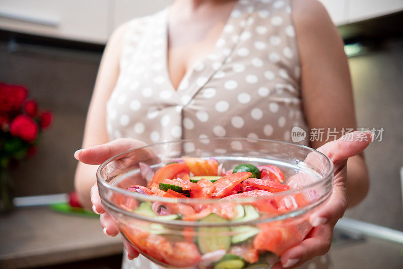 一位身穿棕色连衣裙的年轻女子在米褐色的厨房里烹制素食沙拉。