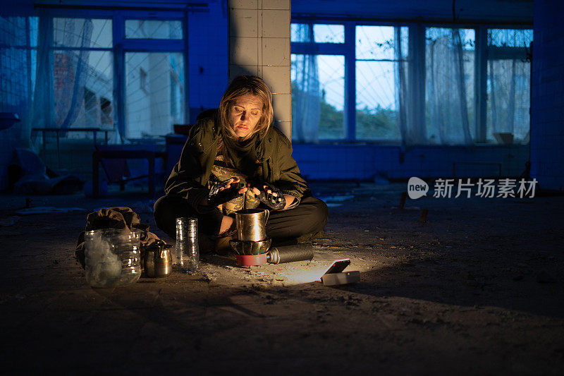 一个又脏又累的女人坐在一间废弃房间的地板上，用煤气炉暖手，为自己准备晚餐。出奇地。在后末日世界徒步旅行。