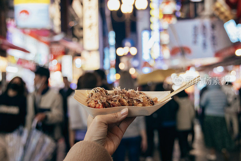 在大阪市中心的街道上，一名女游客正拿着新鲜制作的日本传统街头小吃章鱼烧(章鱼球)。镜头从她的个人角度，我们可以看到她的手拿着滚烫的