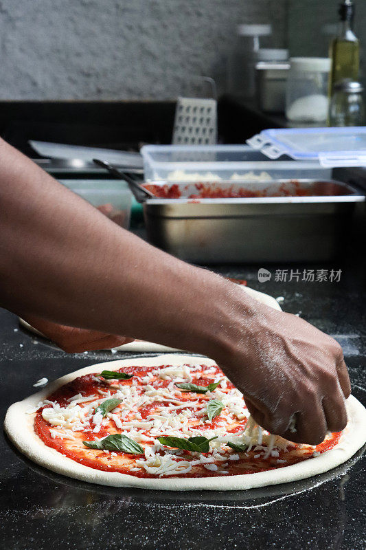 特写图片中，无法辨认的Pizzaiolo(披萨厨师)在未煮熟的披萨基础上添加了撕裂的罗勒叶，玛格丽塔披萨富含番茄马里纳拉酱，磨碎的马苏里拉奶酪，商业厨房的设置，重点放在前景