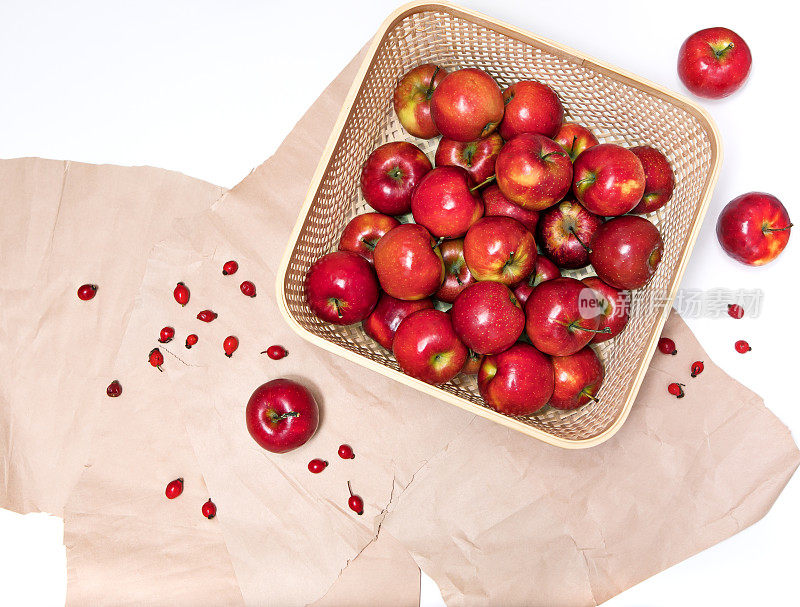丰收的红熟苹果躺在柳条容器里，背景是粗糙的纸和散落的野玫瑰，丰富了身体的维生素