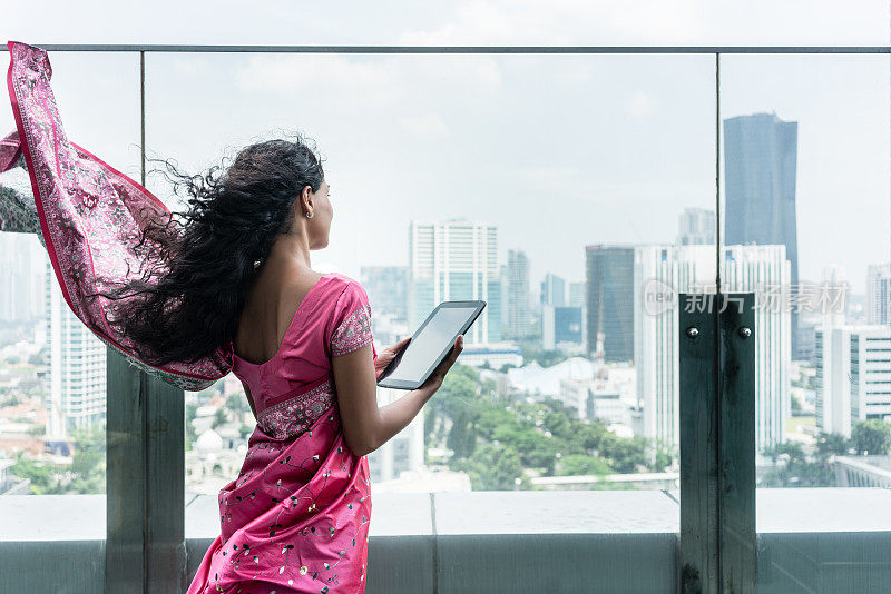 一名年轻的印度女子在一个刮风的日子里在阳台上使用平板电脑