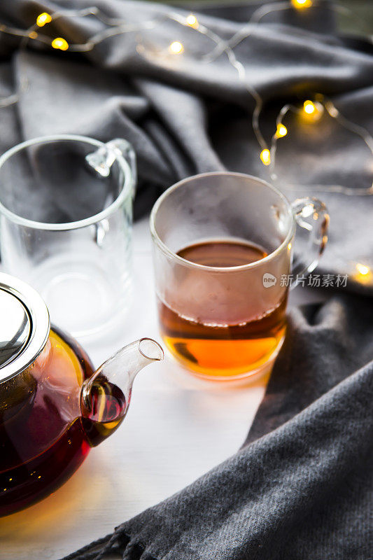 木桌上放着黑色透明茶壶和红茶杯，旁边是一条灰色围巾和灯。冬天家里的概念