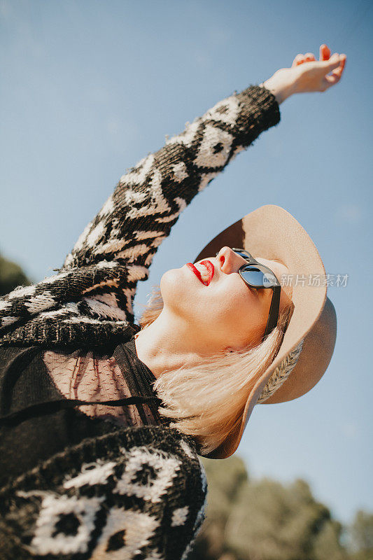 美丽的金发女孩戴着太阳镜，帽子和红唇享受户外，同时举起手臂。