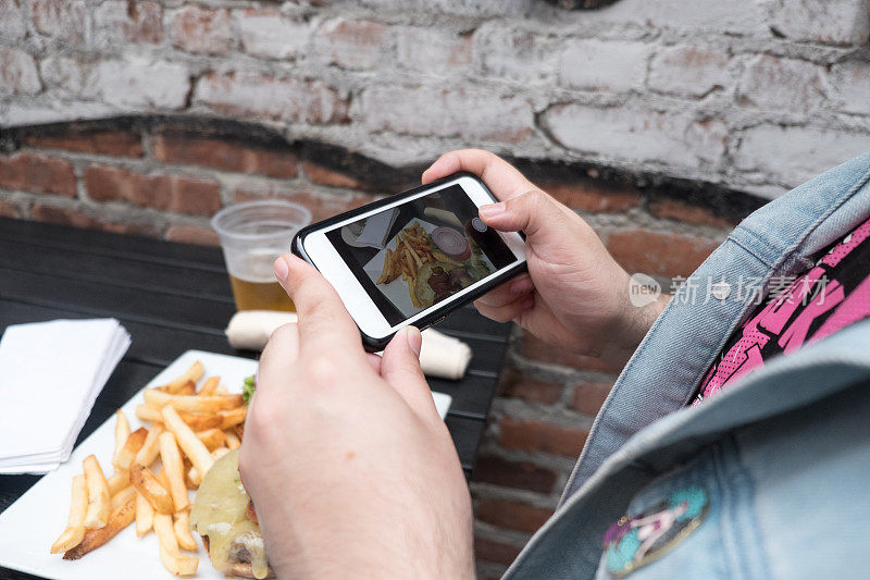 一名男子拿着手机给他的食物拍照。用手机拍摄食物。用智能手机给户外酒吧里的汉堡、啤酒和炸薯条拍照。