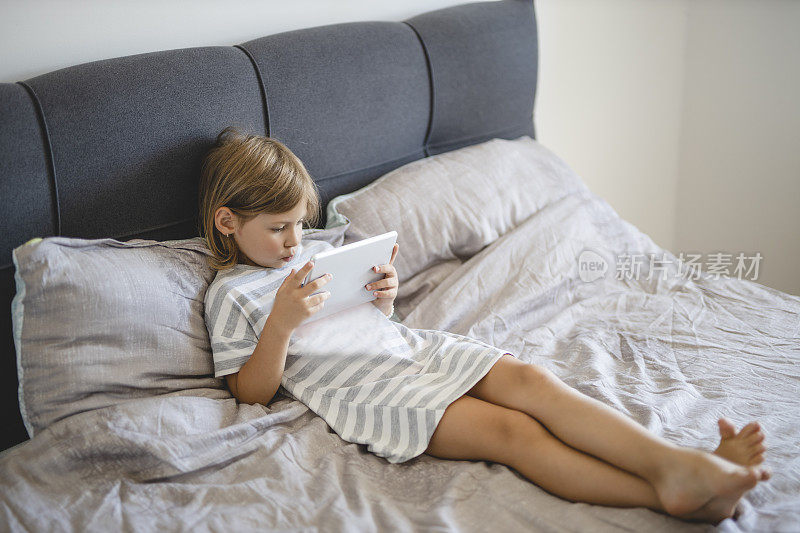 小女孩穿着睡衣，躺在床上，在家里使用电子平板电脑