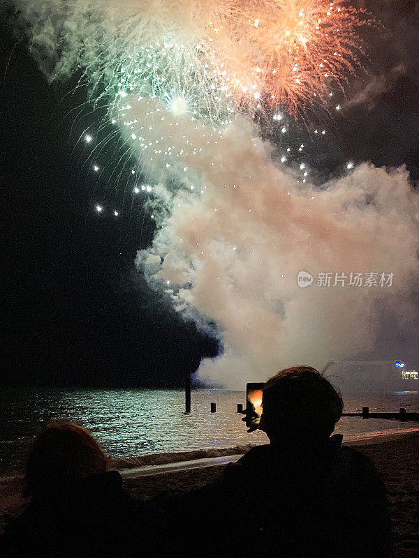 一名年轻女子的剪影特写，她站在海滩的水边，用手机观看并拍摄烟花表演，夜空中的烟雾和火花