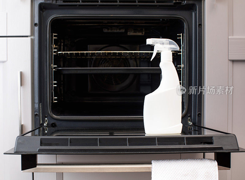 在开放式烤箱的门上有一个白色喷雾瓶的模型，里面装有烤箱清洁剂。厨房的大扫除。
