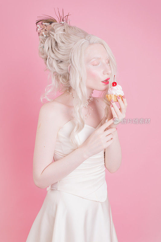 金发女人与美丽的豪华洛可可发型在白色连衣裙粉红色的背景。手里拿着樱桃蛋糕的女孩。复兴。美丽的肖像