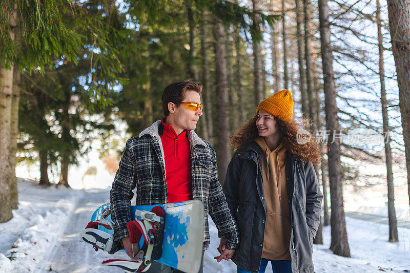 一对美丽的年轻异性恋情侣在雪原森林的小路上散步，手里拿着一块滑雪板