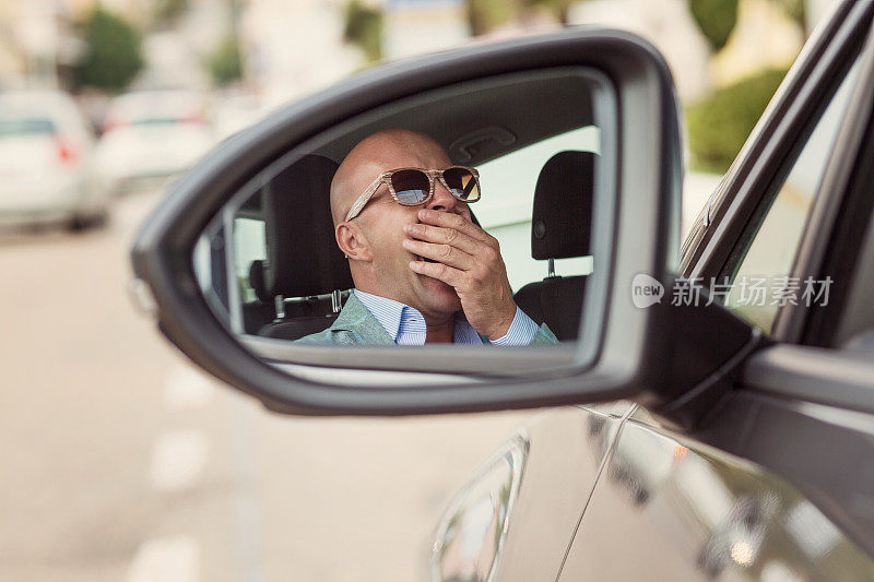 侧镜的视图反射，困倦疲倦，疲劳，打呵欠，疲惫的年轻人戴着太阳镜，驾驶他的汽车在交通经过长时间的驾驶。交通睡眠剥夺事故概念