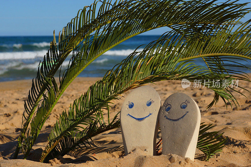 海边的沙滩上，棕榈叶下画着笑脸的沙滩拖鞋