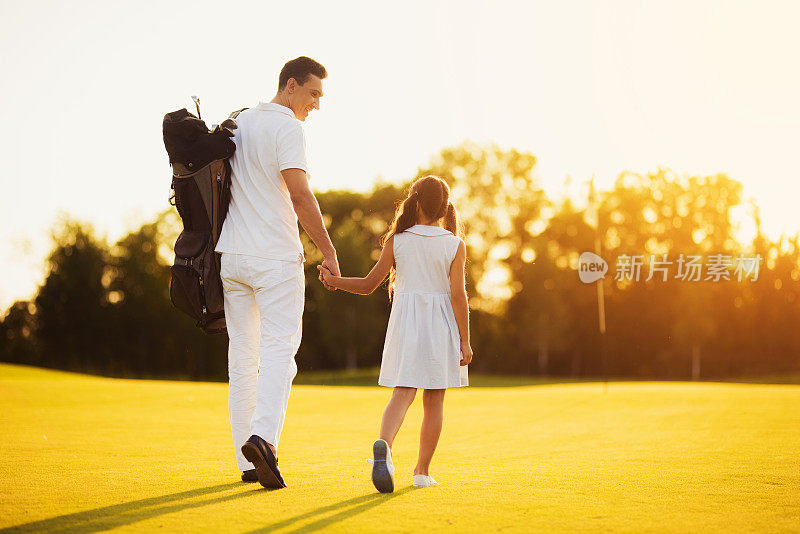 父亲和女儿在夕阳的映衬下走过高尔夫球场。他们背对着镜头