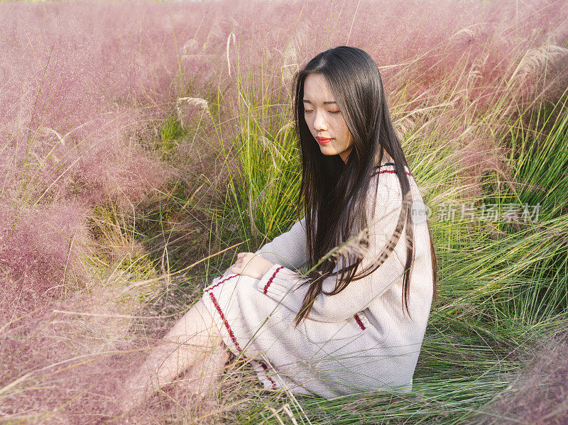 一幅美丽的年轻中国妇女的肖像，穿着白色的毛衣，坐在粉红色的多毛的田野里。